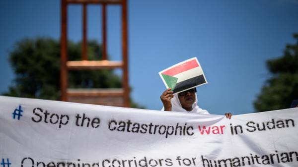 A woman protecting her face with a cardboard bearing the Sudanese flag as she holds a banner next to the mo<em></em>numental wood sculpture 